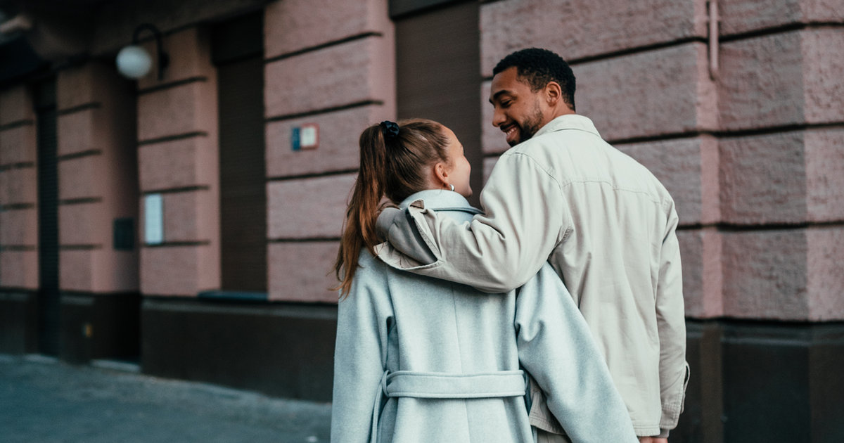 A couple embracing on a city sidewalk beside a brick wall.