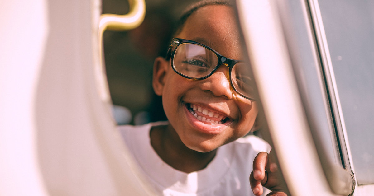A young girl in glasses smiling brightly while leaning out of a car window.